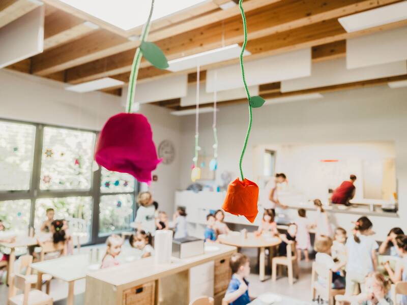 Blick in den Essensraum, der mit Kindern gefüllt ist. Von der Decke hängen zwei bunte Papierrosen