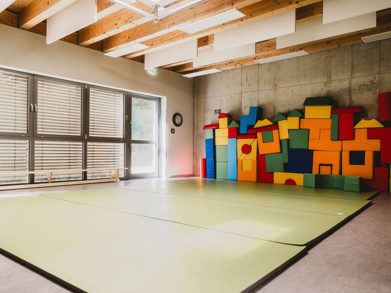 Blick in den Turnraum, auf dem Boden liegen grüne Turnmatten und an der Wand stehen bunte Schaumstoffbausteine