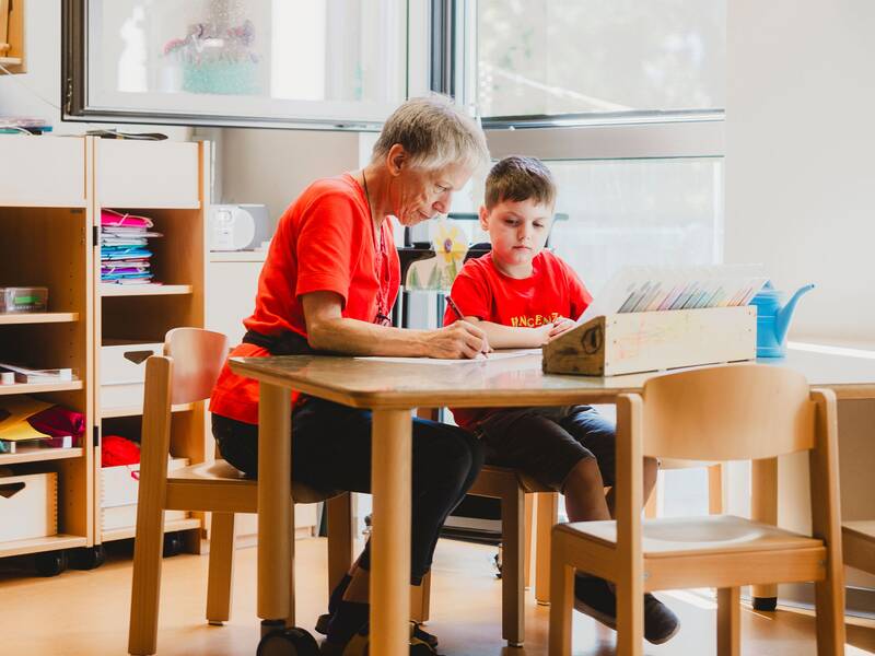 eine Frau im roten T-Shirt sitzt mit einem Jungen an einem Holztisch und hilft ihm beim Malen