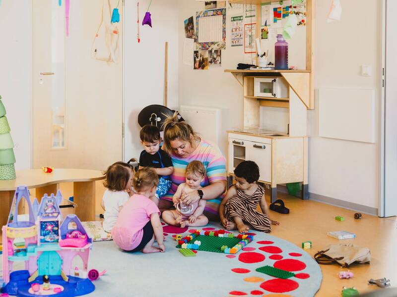 Blick auf eine Erzieherin, die im Gruppenraum mit mehreren Kindern auf einem Spielteppich spielt