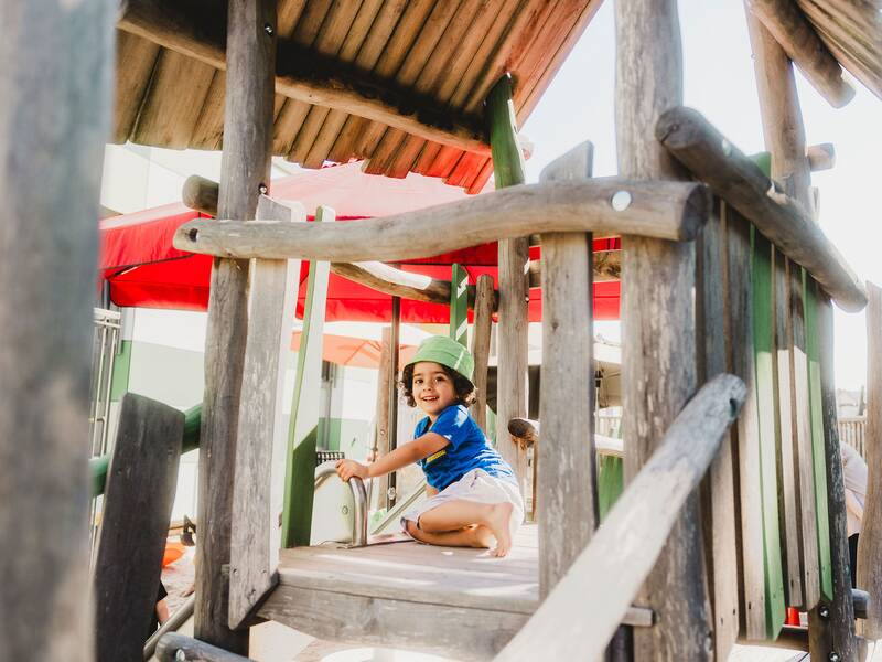 ein Kind sitzt auf einem Holzspielturm auf dem Spielplatz und lächelt in die Kamera