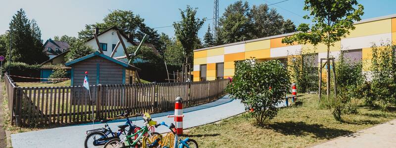 Blick auf den Eingang der Kita, vorne wurden Fahrräder am Fahrradständer abgestellt, dahinter ist eine Wiesenfläche mit Sträuchern. Im Hintergrund sieht man auch den Spielplatz