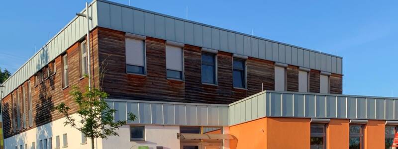 Gebäude der Krippe Pfiffikus Hostenbach. Blauer, strahlender Himmel. Das Gebäude ist zweigeschossig. Die Außenfassade des oberen Stockwerks ist mit Holz verkleidet. Das untere Stockwerk ist weiß gestrichen. Der Vorbau des Gebäudes ist orange gestrichen
