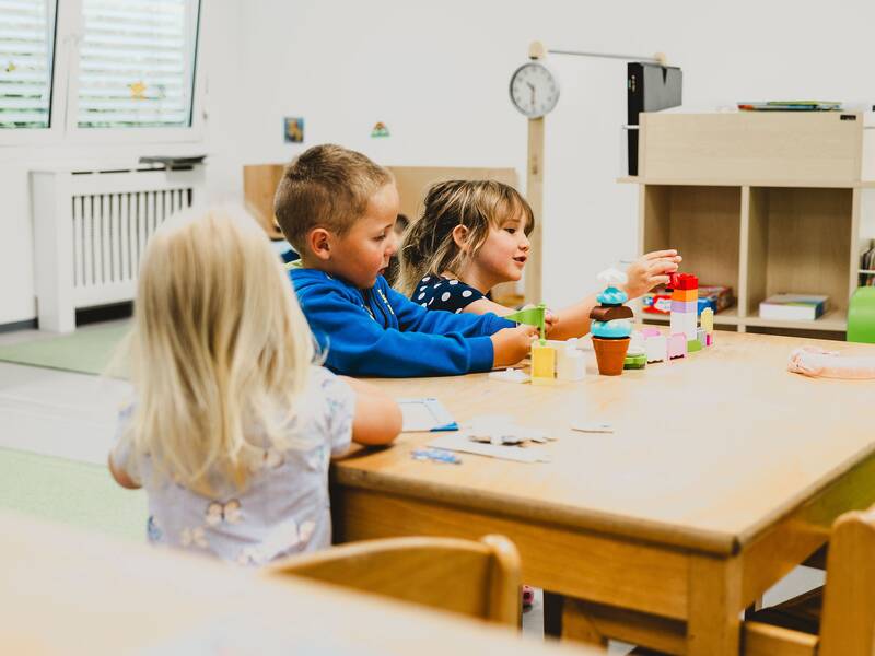 zwei Kinder sitzen an einem Holztisch und bauen mit Legosteinen, ein weiteres Mädchen puzzelt an dem Tisch