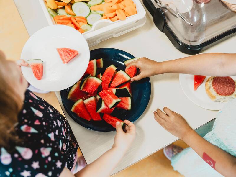 Zwei Kinder nehmen Wassermelonenstücke von einem vollen Teller, während im Hintergrund weitere Snacks sichtbar sind.