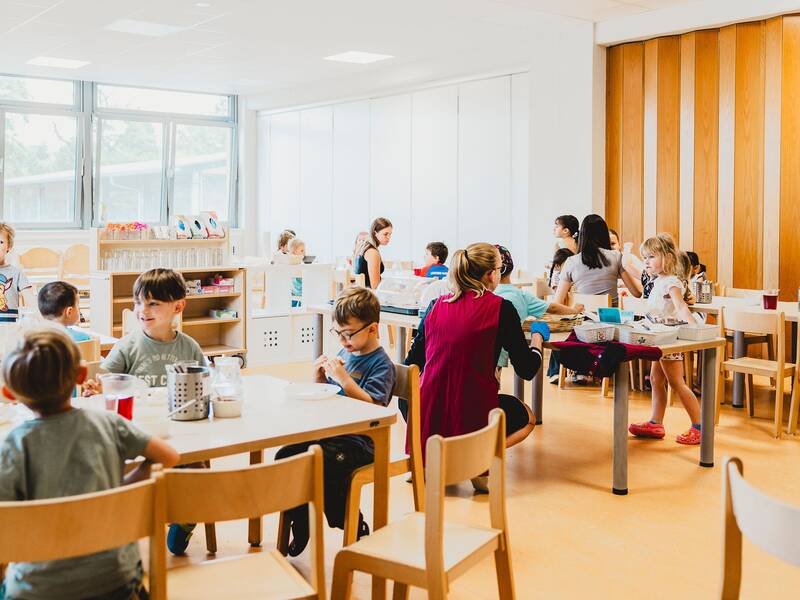 Blick in den Speiseraum in dem die Kinder an den Tischen sitzen und von Erzieherinnen beim Essen begleitet werden