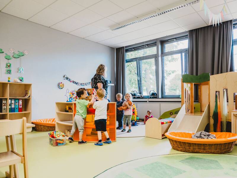 im hellen Gruppenraum spielen mehrere Kinder mit einer Erzieherin.