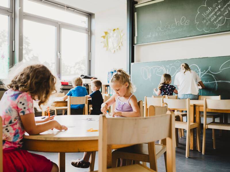 Blick in den Gruppenraum mit einer großen Wandtafel und in dem mehrere Holztische stehen an denen Kinder spielen. Im Vordergrund am Holztisch sitzen zwei Mädchen die ganz vertieft am Malen sind