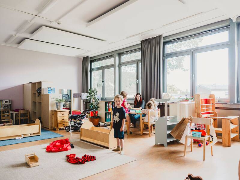 In einem hellen Kindergartenraum spielen mehrere Kinder mit Spielzeug, während eine erwachsene Person am Tisch sitzt.