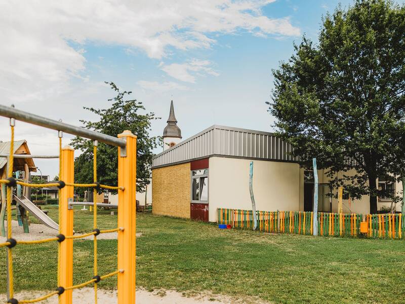 Blick auf den Spielplatz. Zu sehen ist eine Wiese, ein Klettergerüst, ein Spielturm mit Rutsche, ein bunter Holzzaun und im Hintergrund auch noch die Spitze des Kirchturmes