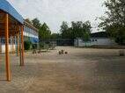 Schulhof der Grundschule im Bisttal, mit Blickrichtung zur Turnhalle. Auf dem Schulhof stehen zwei Tischtennisplatten, zwei Sitzgruppen aus Baumstämmen, ein Sandkasten, der mit einem blauen Sonnensegel überspannt ist.