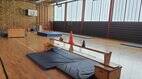 Innenansicht der Turnhalle der Grundschule im Bisttal. Es sind verschiedene Stationen aufgebaut: im Vordergrund eine umgedrehte Bank auf zwei kleinen Kästen, die in einiger Entfernung zueinander stehen. Im Hintergrund: links ein Sprungkasten,  davor Kletterseile, rechts Turnringe.